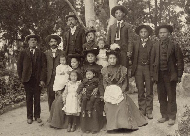 Japanese Family photo with 14 people dressed up with men in suit and ties and women in long dresses. Park setting.