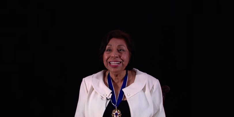 photo of Sylvia Mendez, smiling in white jacket wearing medal around neck.