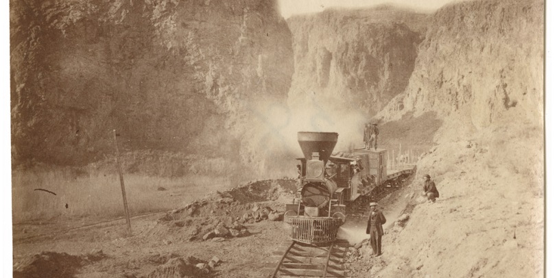 A locomotive passes through a narrow canyon in an old photo.