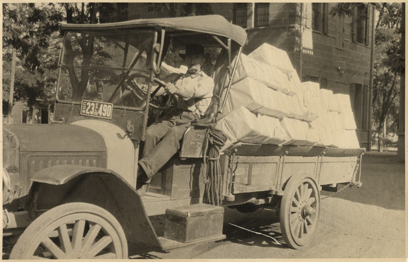 Man driving old truck without doors hauls books wrapped in white paper.