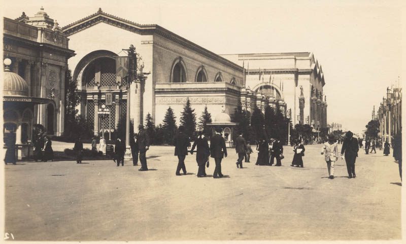 Large white building on the right of a wide walkway with people strolling.