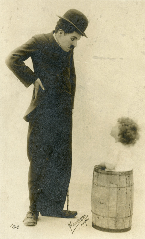 Charlie Chaplin as The Tramp in bowler hat looking down on small child standing in a wood barrel.