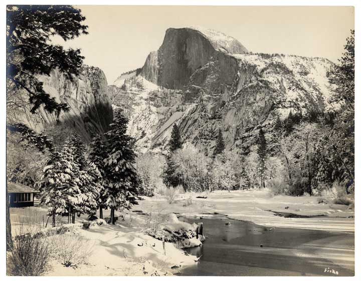 Winter scene of Half Dome from bridge in Yosemite Valley, 1907; shows snow on trees, ground and rock formations; cabin at left, water in foreground.