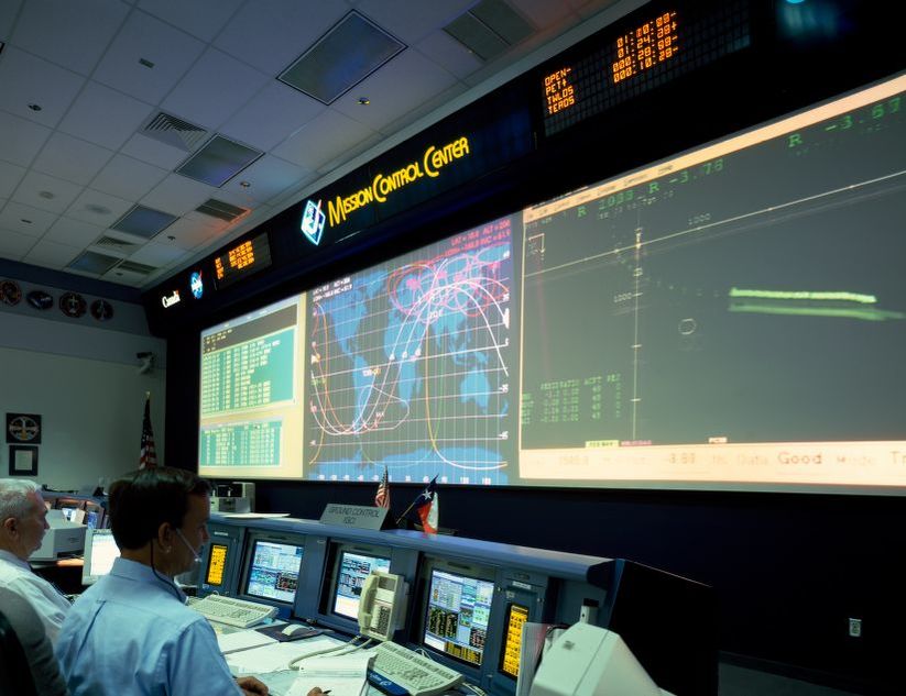 Shows two men in front of a bank of computers and projected screens