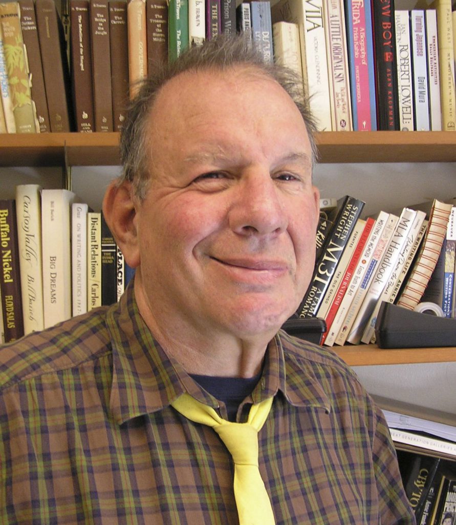 A photograph of a smiling man wearing a plaid shirt and yellow tie, in front of a bookcase.